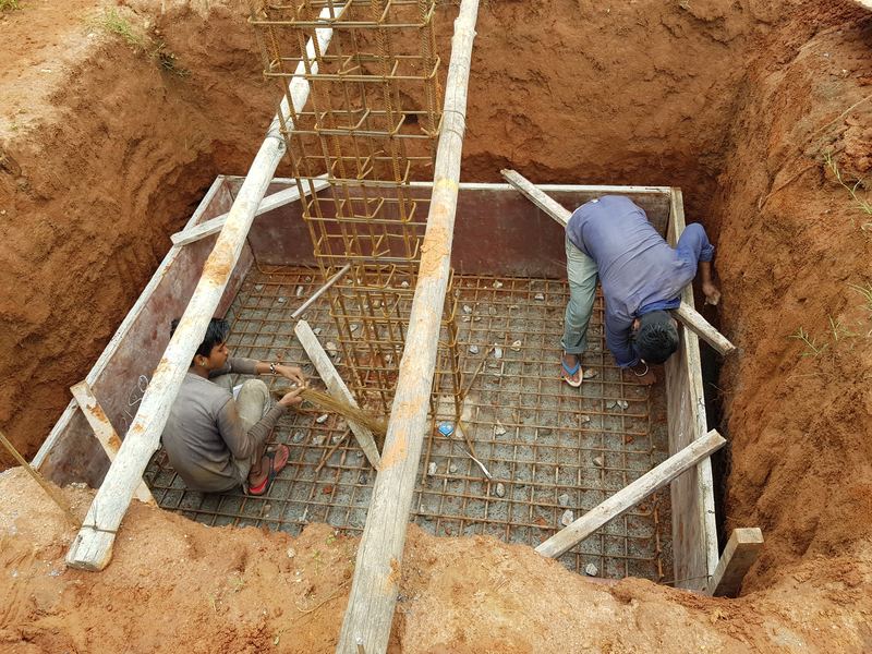 Two workers tying the reinforcement mesh at the bottom of a footing pit in red soil